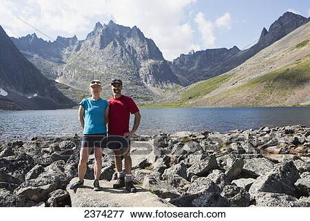 Stock Photo - Couple posing beside a lake in a valley with rugged mountains in the background; Yukon, Canada. Fotosearch