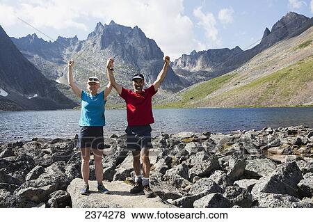 Stock Photo - Couple posing with arms raised beside a lake in a valley with rugged mountains in the background; Yukon, Canada. Fotosearch