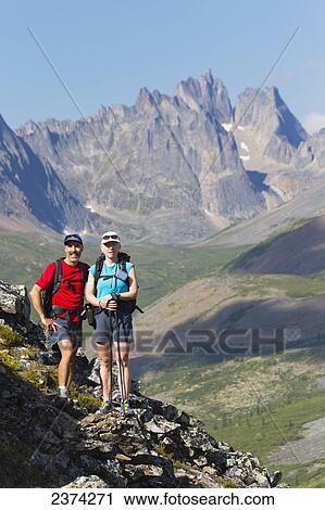 Stock Image - Female and male hiker standing on rock cliff in the mountains with mountain valley and blue sky; Yukon, Canada. Fotosearch