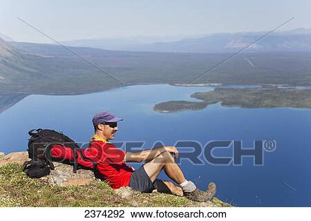Male hiker sitting on mountain side overlooking mountain lake below in the background with blue sky; Yukon, Canada View Large Photo Image Stock Image - Male hiker sitting on mountain side overlooking mountain lake below in the background with blue sky; Yukon, Canada. Fotosearch