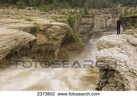 Stock Image - Man watches the Trout River flowing over Whittaker Falls along the Mackenzie Highway; Northwest Territories, Canada. Fotosearch
