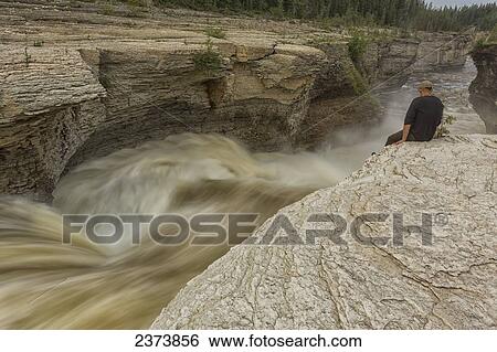 Stock Photograph - Man watches the Trout River flowing over Whittaker Falls along the Mackenzie Highway; Northwest Territories, Canada. Fotosearch