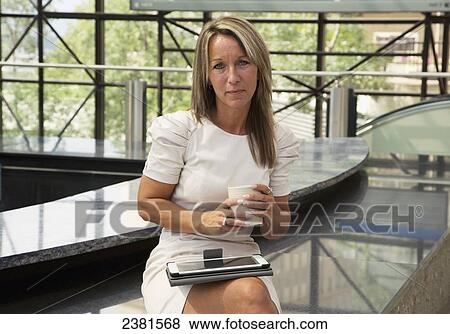 Stock Photo - Mature Professional Businesswoman In Downtown Office Environment Using A Mobile Tablet Device And Drinking A Coffee; Edmonton, Alberta, Canada. Fotosearch
