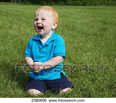 Stock Photograph - Portrait of a baby with red hair sitting in the grass and laughing; Stony Plain, Alberta, Canada. Fotosearch