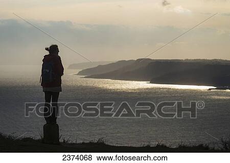 Stock Photo - Silhouette of a woman admiring the view towards Lyme Regis from the top of Golden Cap on the Jurassic Coast; Seatown, Dorset, England. Fotosearch