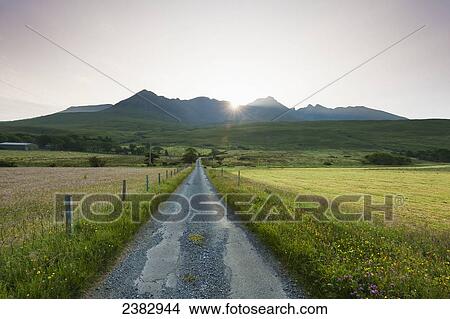 Picture - View along a road early in the morning with the rising sun behind the Black Cuillin ridge; Glen Brittle, Isle of Skye, Scotland. Fotosearch