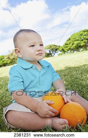 Stock Image - Young boy sitting on the grass at a park with oranges; Honolulu, Oahu, Hawaii, United States of America. Fotosearch