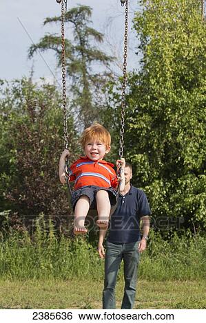 Stock Photograph - A boy swinging on a swing set with his father in the background; Stony Plain, Alberta, Canada. Fotosearch
