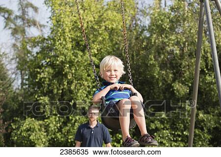 Stock Photography - A boy swinging on a swing set with his father in the background; Stony Plain, Alberta, Canada. Fotosearch