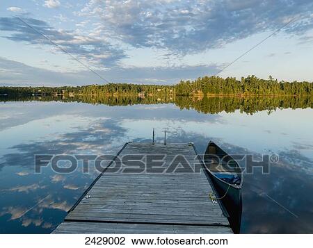 Stock Image - A canoe tied to a wooden dock and clouds reflected in a tranquil lake; Ontario, Canada. Fotosearch