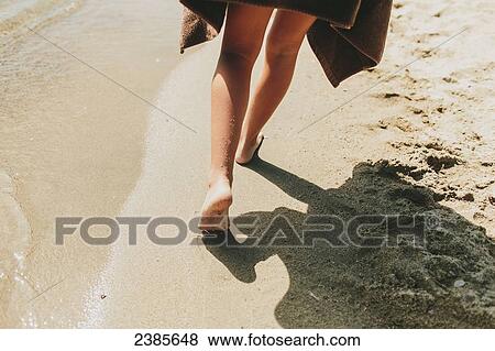 Stock Photo - A girl walks barefoot in the sand along the water's edge; Peachland, British Columbia, Canada. Fotosearch