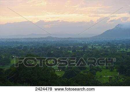 Stock Photo - A lush landscape of trees with mountains in the distance at sunset; Ulpotha, Embogama, Sri Lanka. Fotosearch