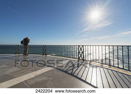 Picture - A man stands taking a photograph at the railing along the water's edge; Nerja, Malaga, Costa del Sol, Andalusia, Spain. Fotosearch