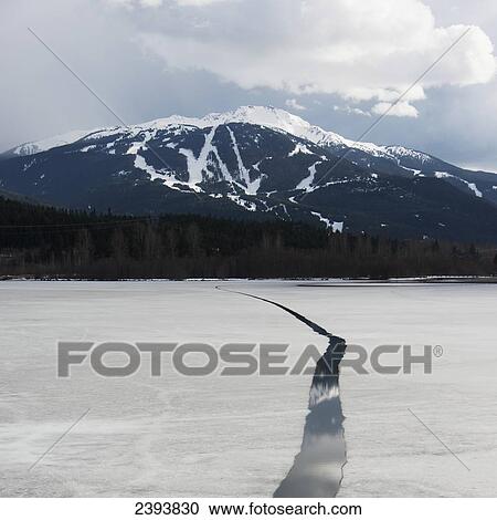 Stock Image - A melted crack in the frozen lake with snowcapped mountains in the distance; Whistler, British Columbia, Canada. Fotosearch