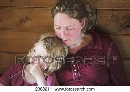 Stock Image - A mother and daughter sitting together reading a book, leaning against a wooden cabin wall; Alaska, United States of America. Fotosearch