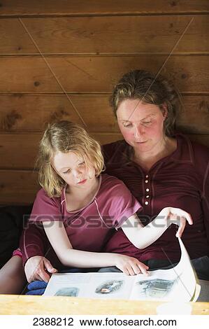 Stock Image - A mother and daughter sitting together reading a book, leaning against a wooden cabin wall; Alaska, United States of America. Fotosearch