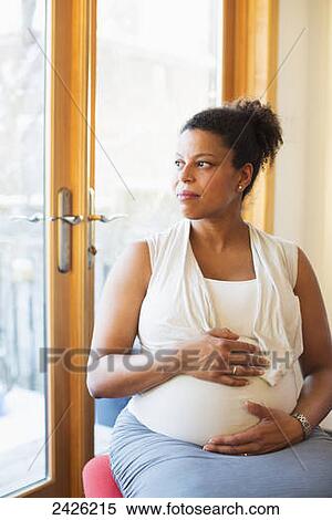 Stock Photography - A pregnant woman with hands on her belly loking out the window; Toronto, Ontario, Canada. Fotosearch
