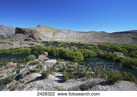 Stock Image - A river lined with lush shrubs and a rock cliff with blue sky; Valle Encatado, Bariloche, Argentina. Fotosearch