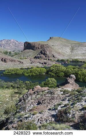Stock Image - A river lined with lush shrubs and a rock cliff with blue sky; Valle Encatado, Bariloche, Argentina. Fotosearch