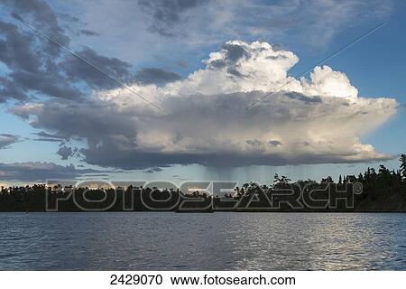 Stock Image - A single storm cloud deposits rain over the silhouette of a forest along the shoreline of a lake; Ontario, Canada. Fotosearch