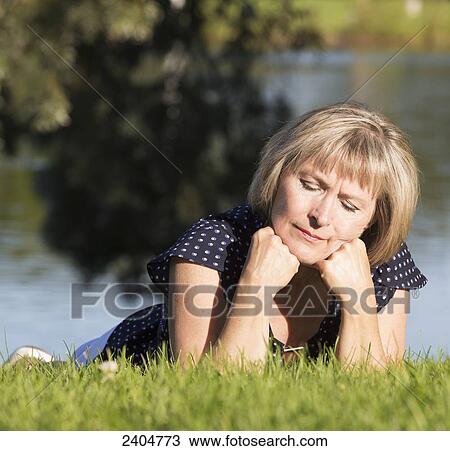 Stock Image - A woman on the grass in a park with a contemplative look on her face; Edmonton, Alberta, Canada. Fotosearch