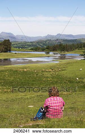 Stock Image - A woman sits on a grassy hill overlooking the landscape; Lake District, Cumbria, England. Fotosearch