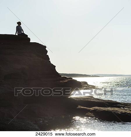 Stock Photography - A woman sits on the top of a rock cliff looking out over the ocean; Green Gables, Prince Edward Island, Canada. Fotosearch