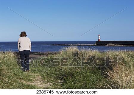Stock Photography - A woman stands on a path at the water's edge with a view of a lighthouse along the coast; Berwick on Tweed, Northumberland, England. Fotosearch