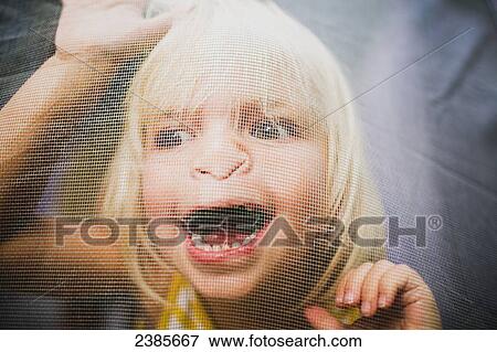 Stock Photo - A young girl with a silly expression on her face viewed through a window screen; Peachland, British Columbia, Canada. Fotosearch