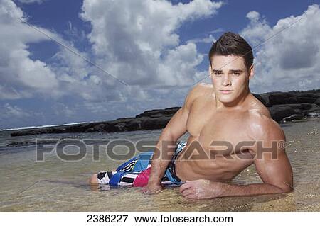 Stock Photo - A young man in a bathing suit lays in the shallow ocean water at the beach; Hawaii, United States of America. Fotosearch