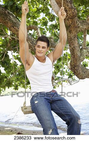 Stock Image - A young man sits on a swing on the beach at the water's edge; Hawaii, United States of America. Fotosearch