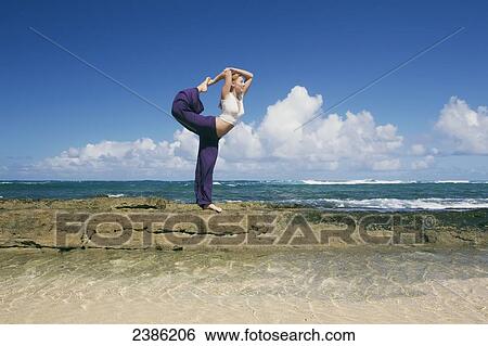 Stock Photograph - A young woman does a yoga pose on a rock wall at the water's edge; Kauai, Hawaii, United States of America. Fotosearch