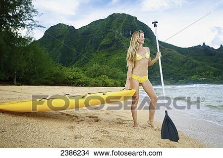 A young woman in a yellow bikini stands at the water's edge with a paddleboard and paddle; Kauai, Hawaii, United States of America View Large Photo Image Picture - A young woman in a yellow bikini stands at the water's edge with a paddleboard and paddle; Kauai, Hawaii, United States of America. Fotosearch