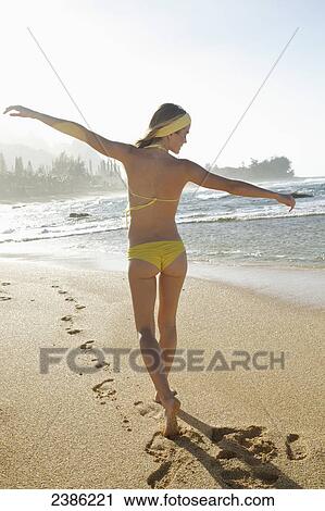 Stock Image - A young woman in a yellow bikini walks on the wet beach along the water's edge; Kauai, Hawaii, United States of America. Fotosearch