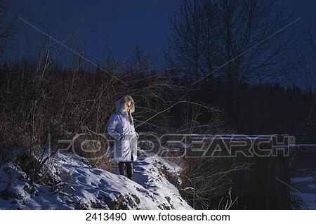 Stock Image - A young woman in winter attire poses at dusk on the riverbank in the snow; Fort McMurray, Alberta, Canada. Fotosearch