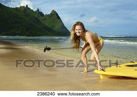 Stock Image - A young woman pulls her stand up paddleboard onto the beach; Kauai, Hawaii, United States of America. Fotosearch