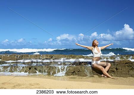 Stock Image - A young woman sits with arms outstretched on a rock wall at the water's edge; Kauai, Hawaii, United States of America. Fotosearch