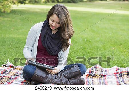 Stock Photo - A young woman sitting in a park using a tablet; Edmonton, Alberta, Canada. Fotosearch