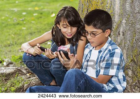 Stock Image - Brother and sister looking at smart phones together in a park; Edmonton, Alberta, Canada. Fotosearch