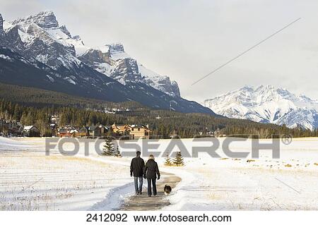 Couple walking a dog on a path in winter with the mountains ahead; Canmore, Alberta, Canada View Large Photo Image Stock Image - Couple walking a dog on a path in winter with the mountains ahead; Canmore, Alberta, Canada. Fotosearch