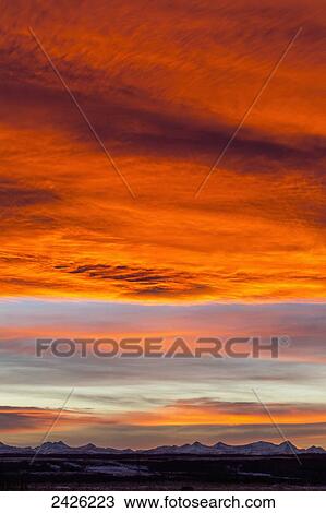Stock Image - Dramatic fiery sunset with orange and red clouds and mountains in the distance; Calgary, Alberta, Canada. Fotosearch