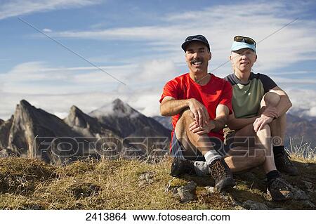Female and male hiking sitting on top of a grassy mountain top with peaks in the distance and blue sky and clouds; Hintertux, Austria View Large Photo Image Picture - Female and male hiking sitting on top of a grassy mountain top with peaks in the distance and blue sky and clouds; Hintertux, Austria. Fotosearch