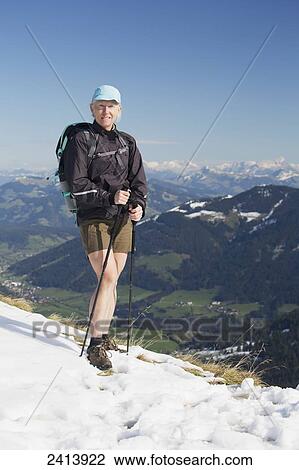 Stock Image - Female hiker on top of snow covered mountain with valley below and mountain ranges in the distance with blue sky; Alpbach, Austria. Fotosearch