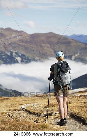 Stock Photograph - Female hiker on trail overlooking a cloud covered valley and mountain range with blue sky and clouds in the distance; Hintertux, Austria. Fotosearch