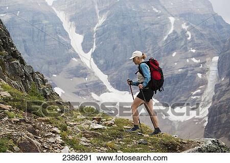Stock Image - Female hiker with poles hiking up mountain path with mountain cliff with snow in background; British Columbia, Canada. Fotosearch