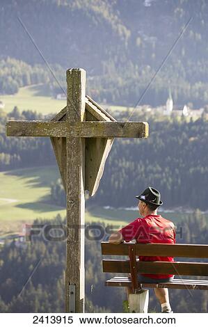 Male hiker sitting on bench with large wooden cross overlooking valley with gren meadows and trees; Aschau, Austria View Large Photo Image Stock Photography - Male hiker sitting on bench with large wooden cross overlooking valley with gren meadows and trees; Aschau, Austria. Fotosearch