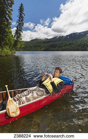 Picture - Man reclined in canoe and checking his camera on Byers Lake with forested foothills in the background, Byers lake campground, Denali State Park; Alaska, United States of America. Fotosearch
