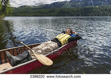 Stock Image - Man reclined in canoe and resting in the sunshine on Byers Lake with forested foothills in the background, Byers lake campground, Denali State Park; Alaska, United States of America. Fotosearch