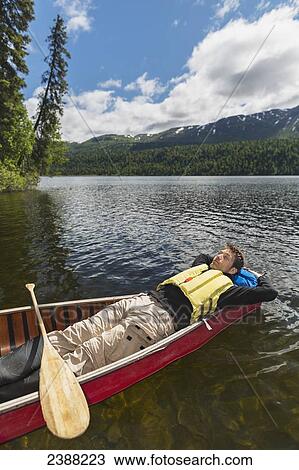 Stock Image - Man reclined in canoe and resting in the sunshine on Byers Lake with forested foothills in the background, Byers lake campground, Denali State Park; Alaska, United States of America. Fotosearch