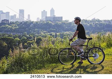 Stock Image - Man riding his electric bicycle with Edmonton city skyline in the background; Edmonton, Alberta, Canada. Fotosearch
